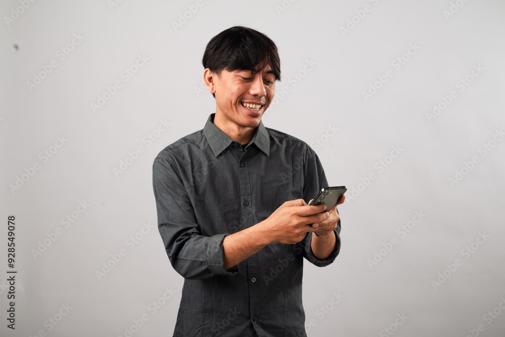 A smiling Asian man in a dark gray shirt interacts with a smartphone, looking down as he types with both hands. The casual and friendly atmosphere suggests he is engaged in a pleasant conversation A smiling Asian man in a dark gray shirt interacts with a smartphone, looking down as he types with both hands. The casual and friendly atmosphere suggests he is engaged in a pleasant conversation