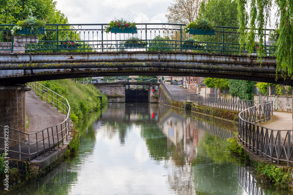 Historic iron bridge over a scenic canal in Montargis, France, with reflections in the calm water and vibrant greenery along the riverside path Historic iron bridge over a scenic canal in Montargis, France, with reflections in the calm water and vibrant greenery along the riverside path