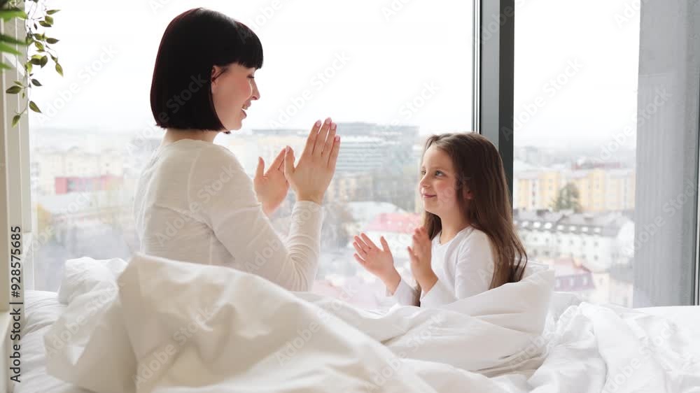 Mother and daughter playing in bed clapping hands. Happy family bonding in morning light. Indoor activity creating joyful moments and connection Mother and daughter playing in bed clapping hands. Happy family bonding in morning light. Indoor activity creating joyful moments and connection