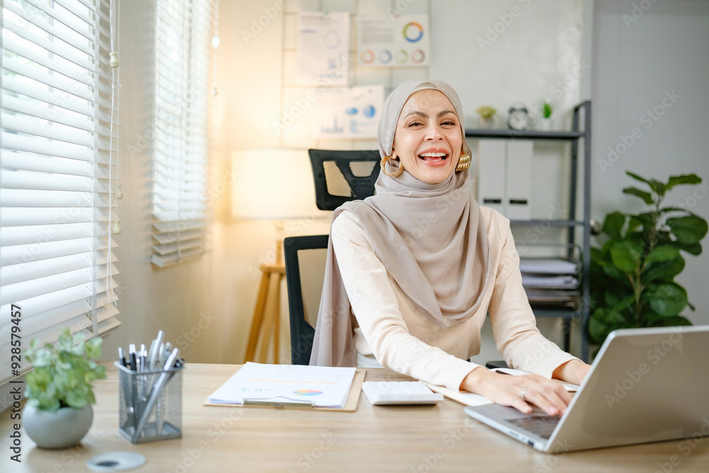 A woman wearing a hijab is sitting at a desk with a laptop and smiling A woman wearing a hijab is sitting at a desk with a laptop and smiling