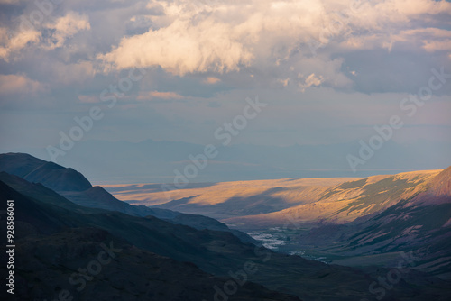 Dramatic aerial top view to golden steppe illuminated by setting sun and mountain range silhouette under clouds in gold sunset tones. Evening alpine landscape under cloudy sky in vivid sunset color.