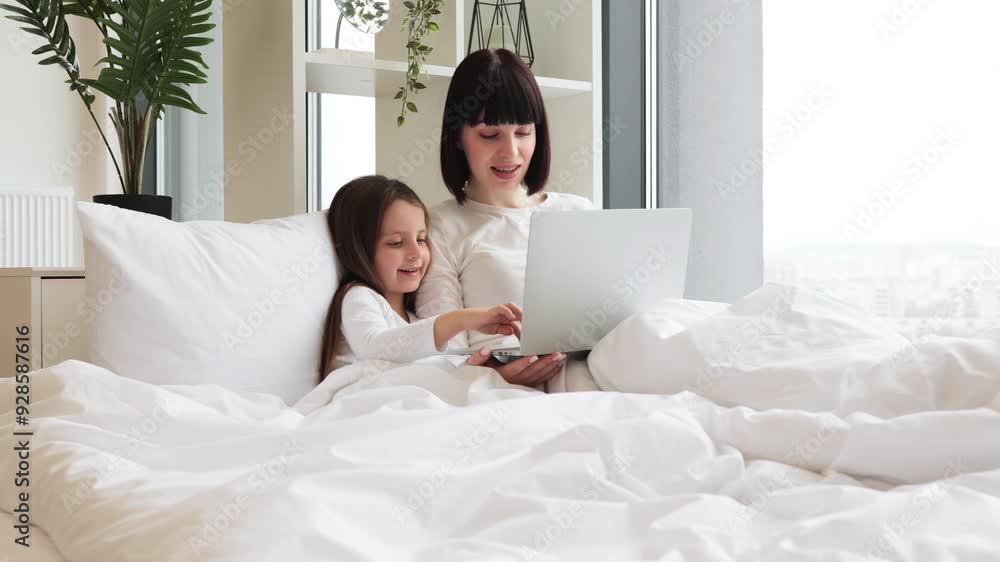 Bright, cozy bedroom creating a warm family atmosphere. Mother and daughter lying in bed using laptop. Bonding time, smiling and enjoying technology. Bright, cozy bedroom creating a warm family atmosphere. Mother and daughter lying in bed using laptop. Bonding time, smiling and enjoying technology.