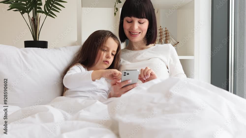 Happy family bonding time, sharing memories and enjoying togetherness in cozy home setting. Mom and daughter lying in bed having fun using smartphone. Happy family bonding time, sharing memories and enjoying togetherness in cozy home setting. Mom and daughter lying in bed having fun using smartphone.