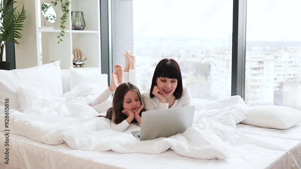 Mother and daughter relaxing in bed using laptop together near large window. Bright and cozy family moment showing love and bonding in comfortable home environment. Mother and daughter relaxing in bed using laptop together near large window. Bright and cozy family moment showing love and bonding in comfortable home environment.