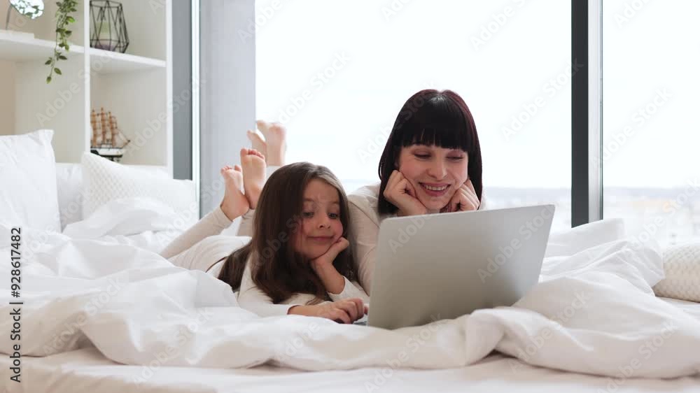 Mother and daughter relaxing in bed using laptop together near large window. Bright and cozy family moment showing love and bonding in comfortable home environment. Mother and daughter relaxing in bed using laptop together near large window. Bright and cozy family moment showing love and bonding in comfortable home environment.
