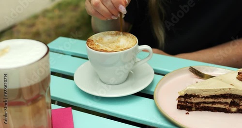 Female vegan stirs cappuccino enjoying rich aroma at table on cafe summer terrace. Woman sits at table with latte made with lactose-free milk and cake