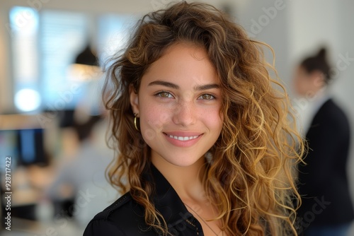 Young woman with curly hair and a bright smile standing in a modern office environment with colleagues working in the background