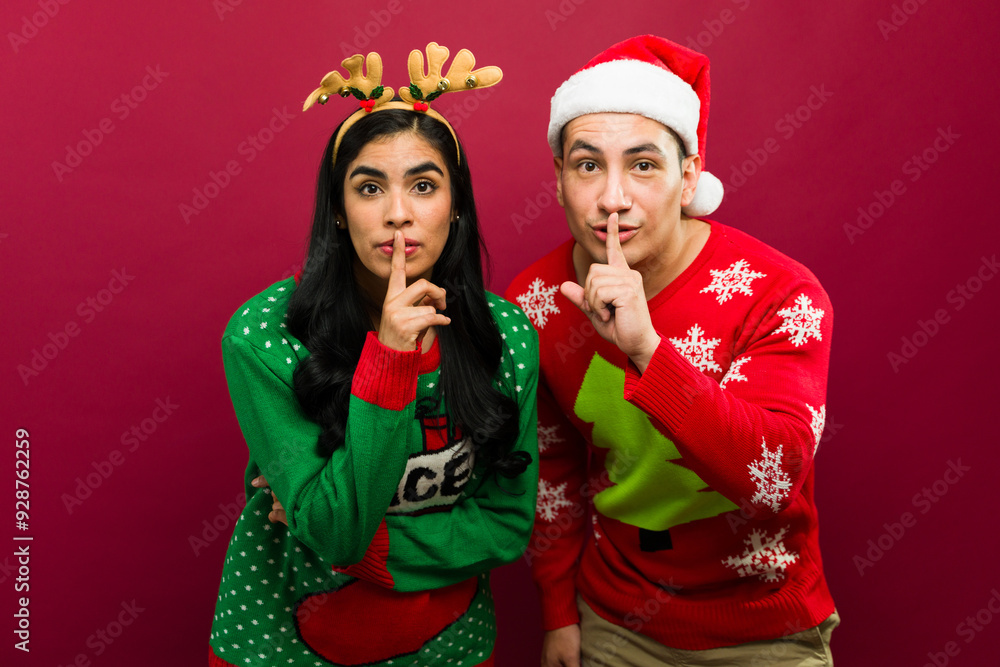 Young couple in ugly sweaters sharing a secret while making a hush gesture against a red backdrop Young couple in ugly sweaters sharing a secret while making a hush gesture against a red backdrop