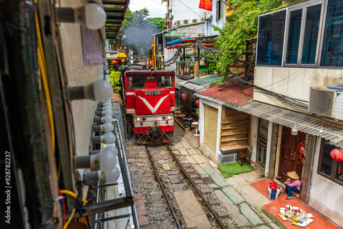 A train passes through a narrow street in Hanoi's Old Quarter. Tourists take photos of the speeding train. Hanoi's Train Street is a popular tourist attraction.