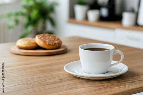 A white ceramic cup filled with black coffee on a saucer, placed on a wooden table with blurred donuts and greenery in the background.