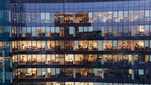 The drone flies high above a glass skyscraper, its windows glowing with light from offices where dedicated employees are working late, showcasing the intensity of urban business life.