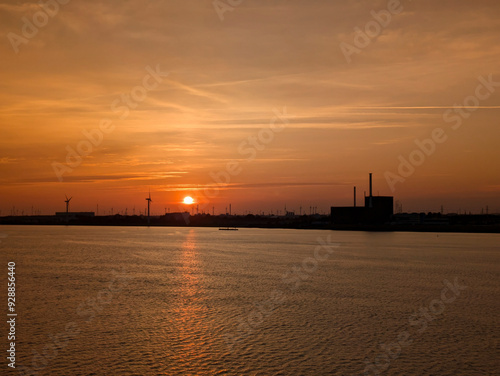 Ein stimmungsvoller Sonnenuntergang über einer Wasserfläche, an deren Horizont die Silhouetten von Windkrafträdern und Industrieanlagen vor einem warmen, orangefarbenen Himmel prangen.