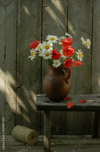 Floral still life with a bouquet of poppies and daisies in a brown clay jug on a wooden bench against a wall of boards photo