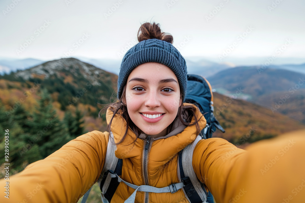 Naklejka premium Joyful hiker capturing a moment in autumn mountains, wearing a warm yellow jacket and gray beanie, surrounded by vivid foliage and distant peaks under a cloudy sky