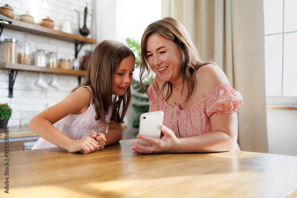 Cheerful mother and daughter using smartphone together in kitchen Cheerful mother and daughter using smartphone together in kitchen