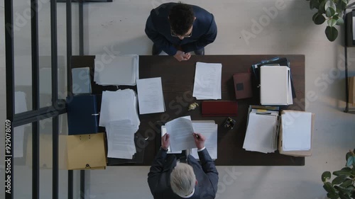 Top down view of two colleagues in office, male assistant standing in front of his boss and handing him documents, furious senior boss tearing them apart and throwing on desk photo