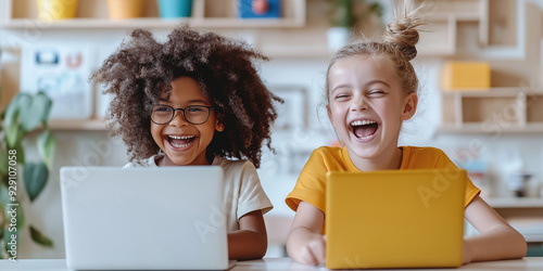 Joyful Moments: Multiethnic Kids Engaged in Learning. Two cheerful primary school kids, a girl with curly hair and glasses and a girl with a bun, laugh together while using laptops in a bright