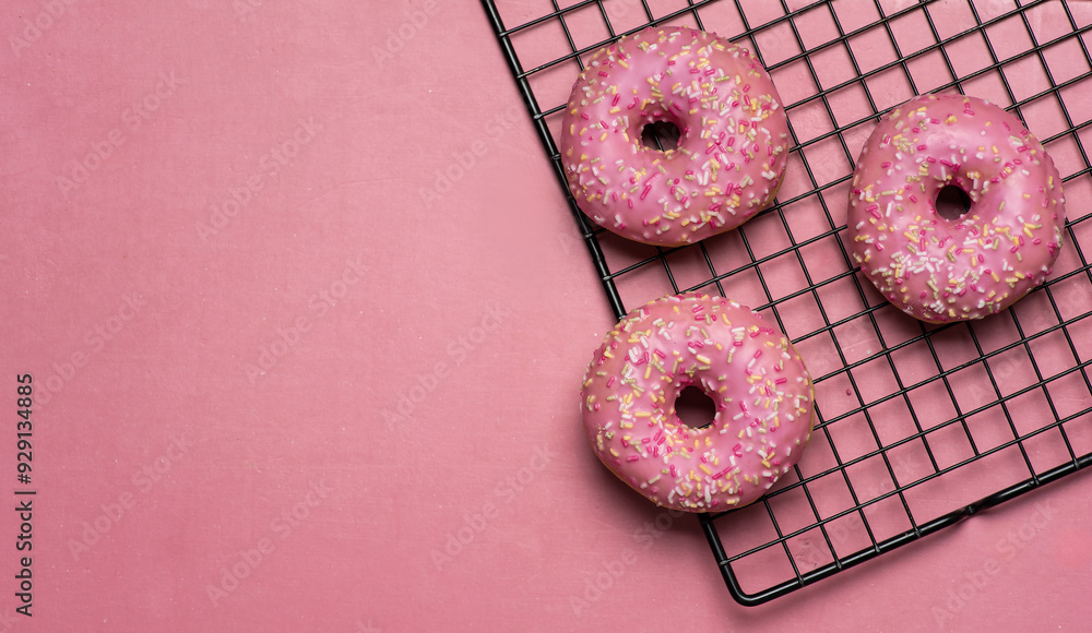 Trio of pink sprinkled donuts partially on cooling rack with pink background.