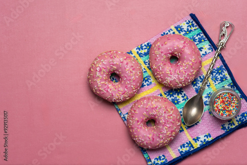 Three pink frosted sprinkle donuts displayed with spoon and sprinkles jar. photo