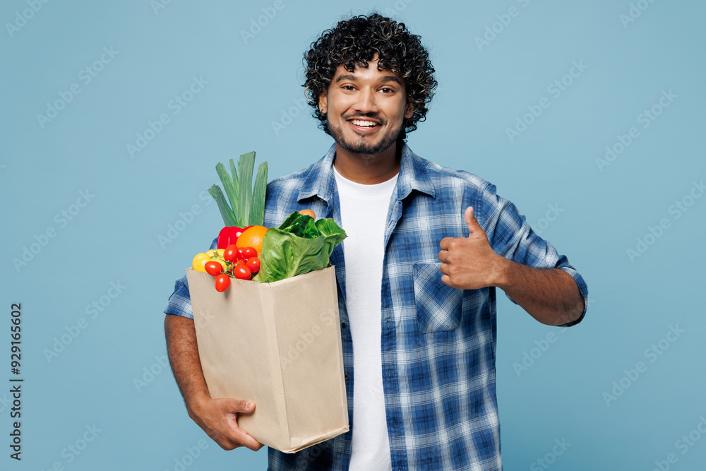 Young smiling happy Indian man wear shirt hold brown craft bag for takeaway mock up with food products show thumb up gesture isolated on plain blue background Delivery service from shop or restaurant Young smiling happy Indian man wear shirt hold brown craft bag for takeaway mock up with food products show thumb up gesture isolated on plain blue background Delivery service from shop or restaurant