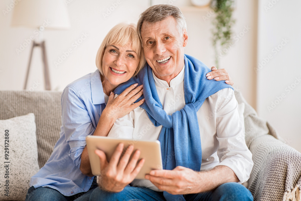 Excited mature couple with digital tablet smiling to camera, woman embracing husband Excited mature couple with digital tablet smiling to camera, woman embracing husband
