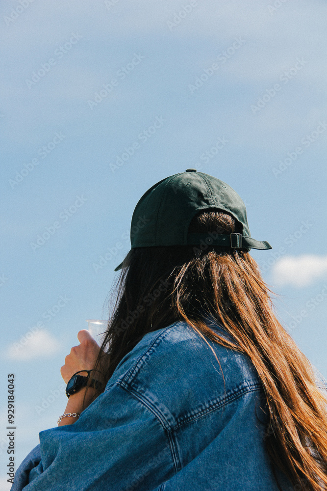 A young woman with long hair, seen from behind, wears a green cap and denim jacket. The soft focus and subtle graininess add an artistic depth to this contemplative scene.