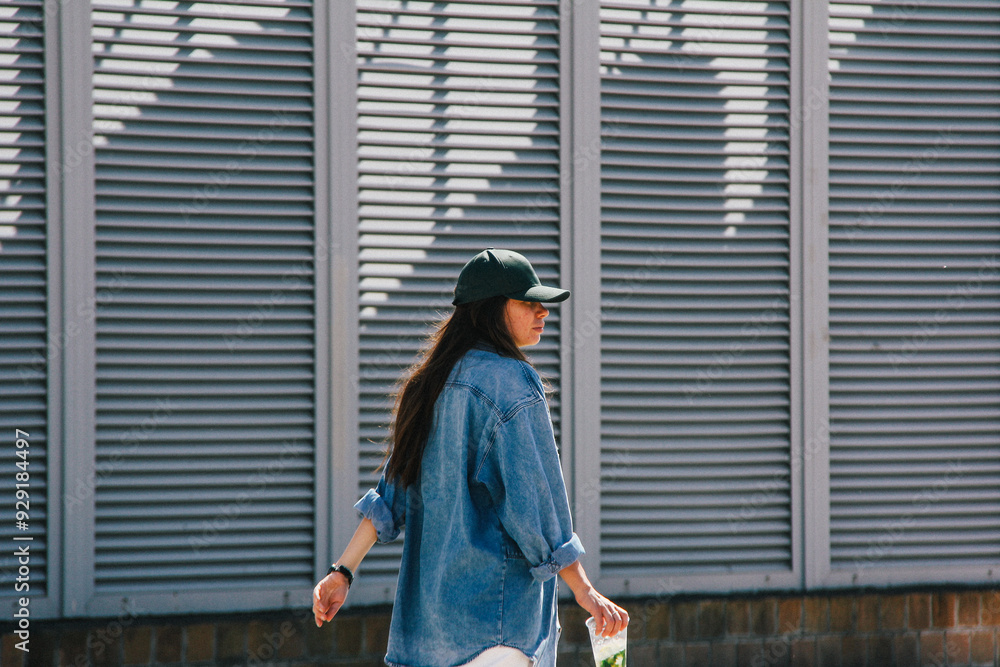 A woman in a casual outfit walks outdoors, her denim jacket and green cap blending with the urban backdrop. The slight blur and natural grain give the image a relaxed, artistic feel.