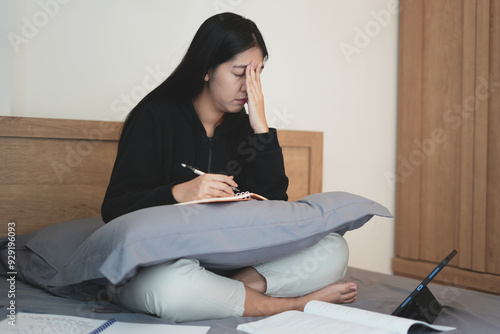 A young asian teenager struggle doing homework with tablet and notebook white sitting on bed, learning at home and getting headache. people struggle with technology and try hard to pass the exam.