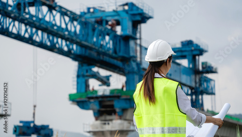 Construction workers wearing a yellow vest and a white helmet is standing in front of a large blue crane.