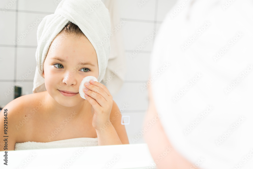 Portrait of a beautiful little girl looking at herself in the mirror cleansing her face with lotion. A pre-adolescent girl or teenager holds a cotton pad in her hands while caring for her skin. Portrait of a beautiful little girl looking at herself in the mirror cleansing her face with lotion. A pre-adolescent girl or teenager holds a cotton pad in her hands while caring for her skin.
