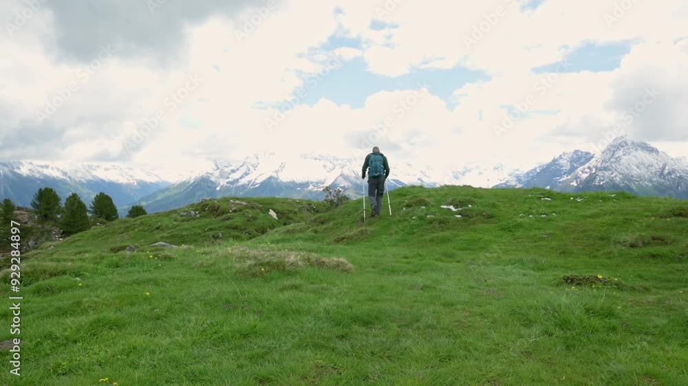 A hiker walks along a green mountain trail with trekking poles, surrounded by lush meadows and overlooking snow-capped peaks of the Alps under a partly cloudy sky. High quality 4k footage A hiker walks along a green mountain trail with trekking poles, surrounded by lush meadows and overlooking snow-capped peaks of the Alps under a partly cloudy sky. High quality 4k footage