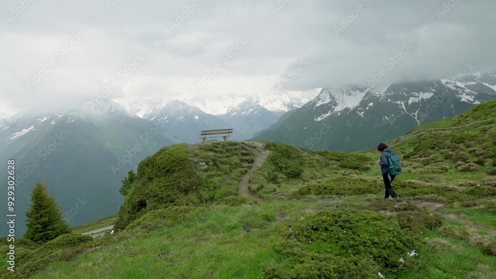 A hiker walks along a scenic mountain path in the Alps, with snow-capped peaks in the background and a bench offering tranquil resting spot, surrounded by lush greenery and dramatic clouds. 4k footage A hiker walks along a scenic mountain path in the Alps, with snow-capped peaks in the background and a bench offering tranquil resting spot, surrounded by lush greenery and dramatic clouds. 4k footage