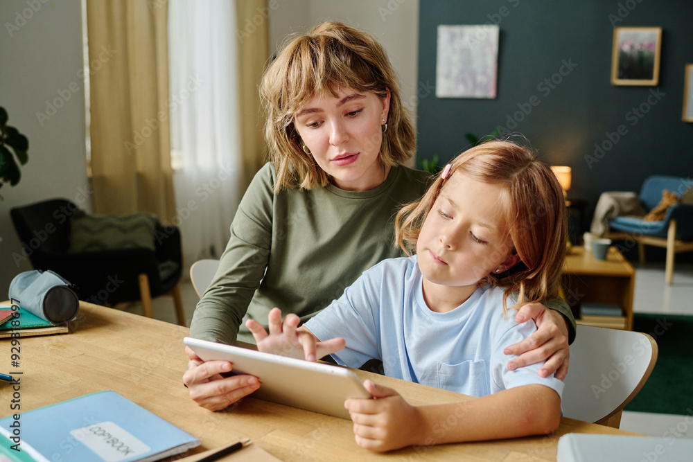 Young Caucasian mom sitting at table with her daughter and hugging her while girl drawing via tablet Young Caucasian mom sitting at table with her daughter and hugging her while girl drawing via tablet
