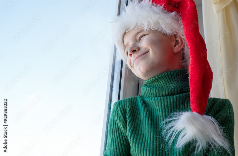 Children's dreams concept. Friendship. A lonely little boy in a Santa Claus hat is sad at the window waiting for gifts, looking out the window, the child is waiting for Santa Claus. The baby is