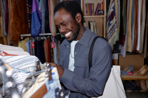 Portrait shot of young smiling African American man in gray trendy shirt choosing clothes shopping at second hand store, camera flash, copy space photo