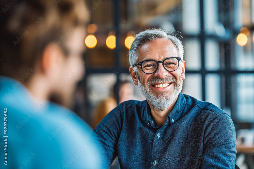 A mature businessman smiles warmly during a meeting with a younger colleague, sharing his expertise and experience in a modern office setting. A mature businessman smiles warmly during a meeting with a younger colleague, sharing his expertise and experience in a modern office setting.
