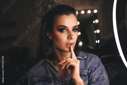 Fotografie A close-up of a perfectly made-up brunette with smoky eyes in the studio, with a backlit mirror behind her on a brown background