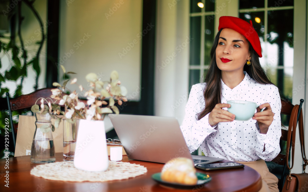 Stylish beautiful smiling cheerful brunette woman in a red beret resting and having croissant breakfast with coffee in city cafe sitting at table on the terrace with phone and laptop on the table Stylish beautiful smiling cheerful brunette woman in a red beret resting and having croissant breakfast with coffee in city cafe sitting at table on the terrace with phone and laptop on the table