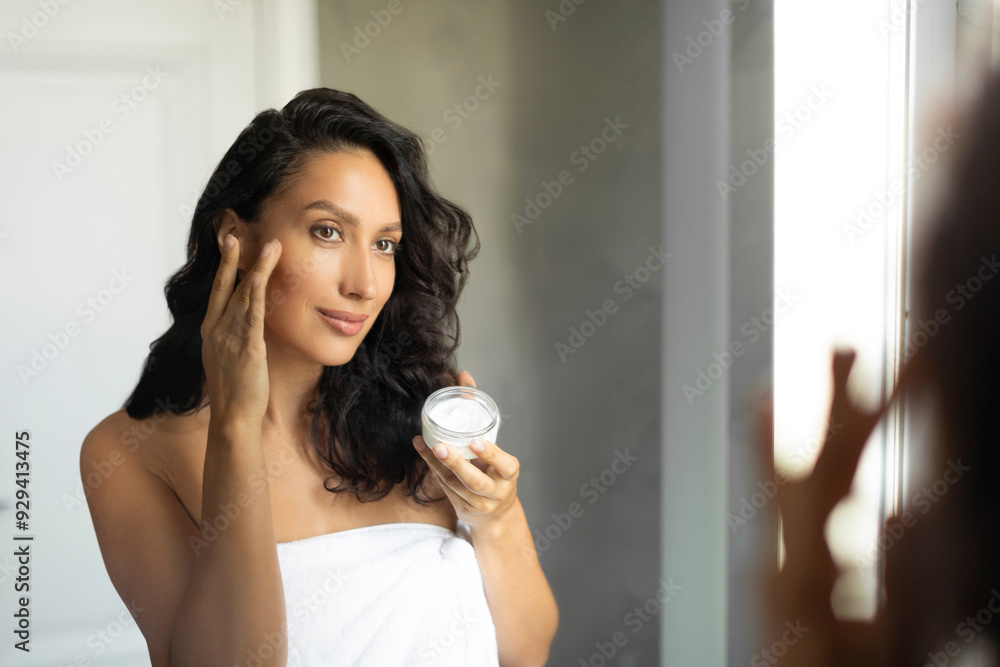 Young European woman applying nourishing cream on face, standing wrapped in towel, holding jar with new anti-aging cosmetics product Young European woman applying nourishing cream on face, standing wrapped in towel, holding jar with new anti-aging cosmetics product