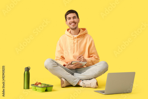 Young man with notebook and lunchbox sitting on yellow background