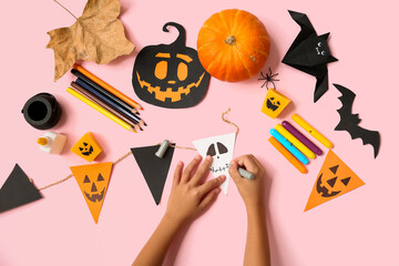 Child with supplies and decorations coloring Halloween garland of flags on pink background
