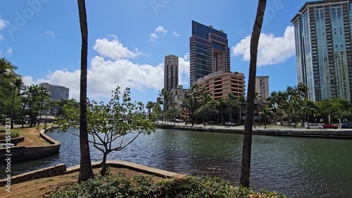 panning footage of the Kalakaua Avenue Bridge over the Ala Wai Canal with skyscrapers hotels,, lush green palm trees, rippling water, blue sky and clouds in Honolulu Hawaii USA photo