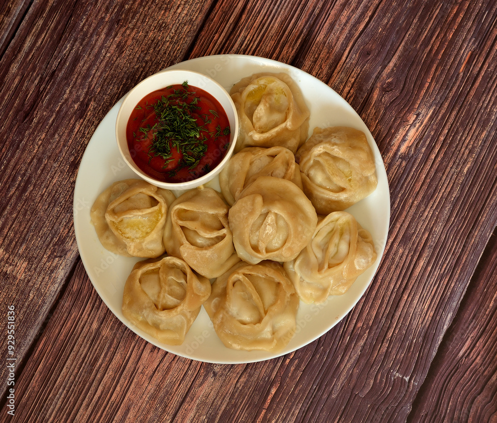 Freshly cooked juicy manti on a large round plate and a cup with tomato sauce and herbs on a wooden table. Freshly cooked juicy manti on a large round plate and a cup with tomato sauce and herbs on a wooden table.