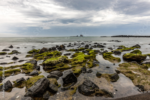 Andeok-myeon, Seogwipo-si, Jeju-do, South Korea - March 13, 2022: Morning view of green laver on volcanic rocks at Sagye Beach against Hyeongjeseom Island on sea horizon photo