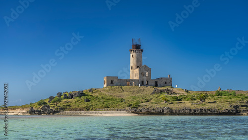 An old dilapidated lighthouse on an island in the ocean. A tall tower against a clear blue sky. The green grass on the hill. Turquoise sea water in the foreground. Mauritius. Île au Phare 