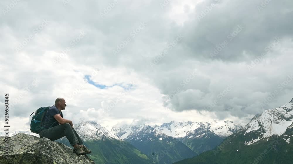 A bald hiker with a backpack sits on a rock, taking in the expansive view of snow-capped mountains under a cloudy sky. Perfect for themes of adventure, travel, and solitude in nature. 4k footage A bald hiker with a backpack sits on a rock, taking in the expansive view of snow-capped mountains under a cloudy sky. Perfect for themes of adventure, travel, and solitude in nature. 4k footage