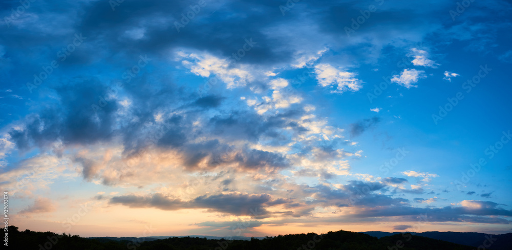 Fototapeta premium Breathtaking sky filled with dramatic clouds transitions from deep blue to soft pastels as sun sets. Horizon lined with silhouetted hills and forests, adding depth to serene and expansive landscape.