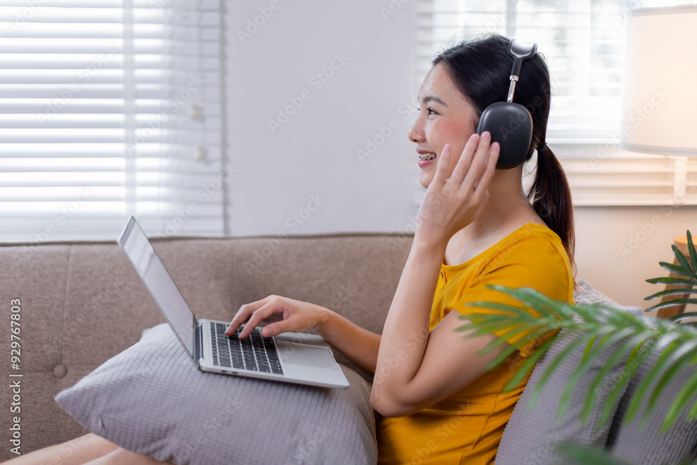 Shot of beautiful young asian woman working with her laptop while listening to music with earphones sitting on a couch at home. Shot of beautiful young asian woman working with her laptop while listening to music with earphones sitting on a couch at home.