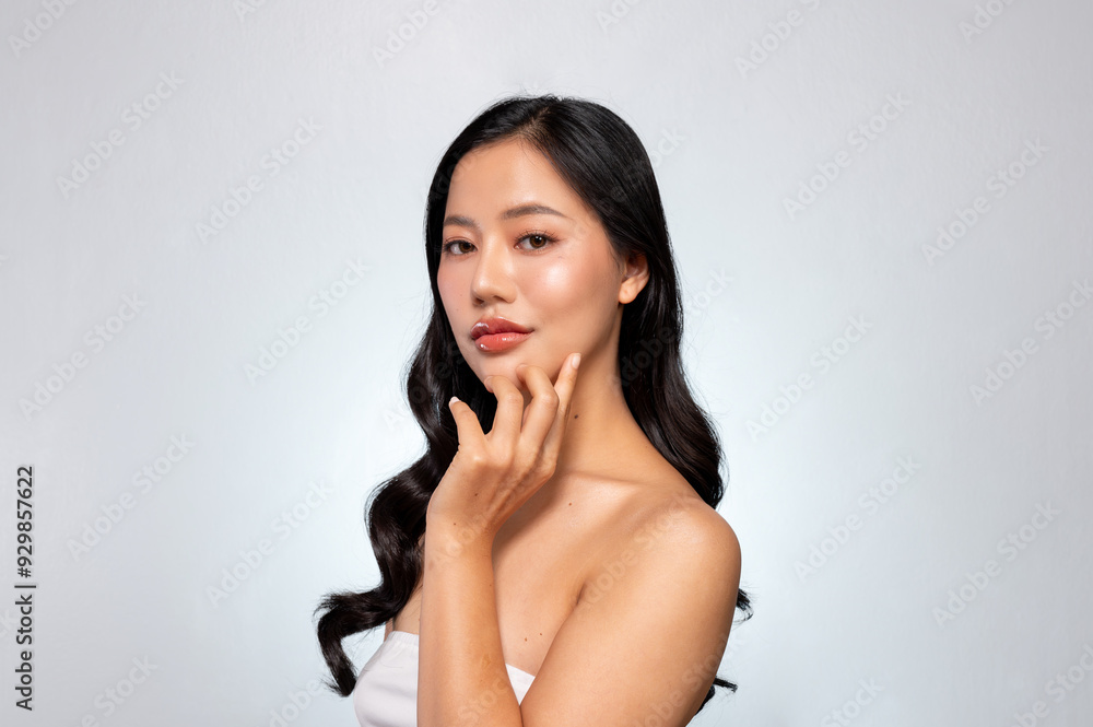 Obraz premium Elegant Asian woman poses against a grey background, looking at the camera while touching her chin.