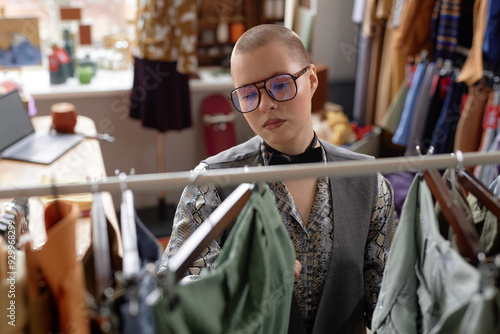 Portrait shot through clothing rack of fashionable female customer in vintage outfit and trendy aviator glasses choosing second hand clothes at thrift store, copy space photo